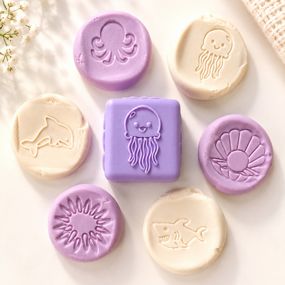 A set of colorful sea-themed soap bars arranged on a light cream background with decorative dried flowers and a woven fabric corner. The soaps include a lavender rectangular bar with a smiling octopus, several round lavender soaps with embossed sea creatures like an octopus, jellyfish, shell, sun, shark, and whale, and several cream-colored soaps with embossed sea creatures.