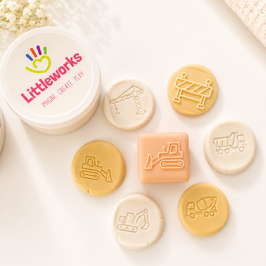 A peach-colored silicone baby teethers or sensory blocks with embossed construction vehicle designs, displayed on a light beige surface with soft shadows and baby's breath flowers.