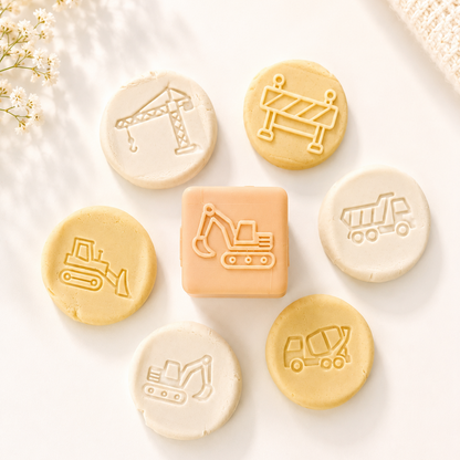 A set of construction-themed baby teethers or bath toys arranged on a white surface. The set includes a central square piece with an embossed excavator, surrounded by six round pieces with embossed construction icons: a crane, a barrier, a bulldozer, a dump truck, a excavator, and a dump truck.